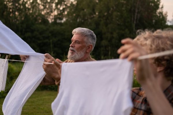Elderly man and teenage boy working together, hanging laundry in a green outdoor setting.