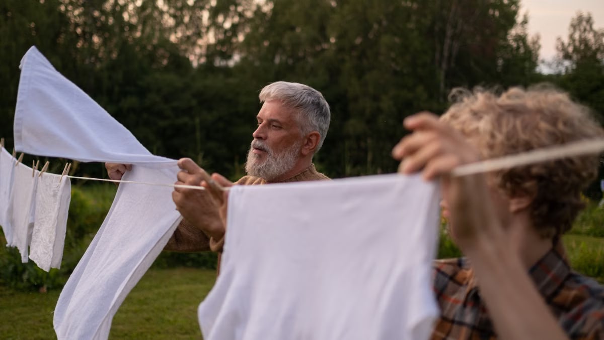 Elderly man and teenage boy working together, hanging laundry in a green outdoor setting.